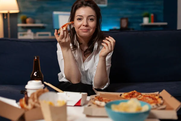 Портрет усміхненої жінки після офісної роботи, що їсть шматочок гарячої доставки піци під час перегляду телевізора — стокове фото