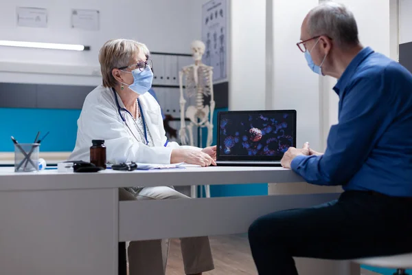 Physician explaining coronavirus bacteria on laptop to senior man, wearing face mask — Stockfoto