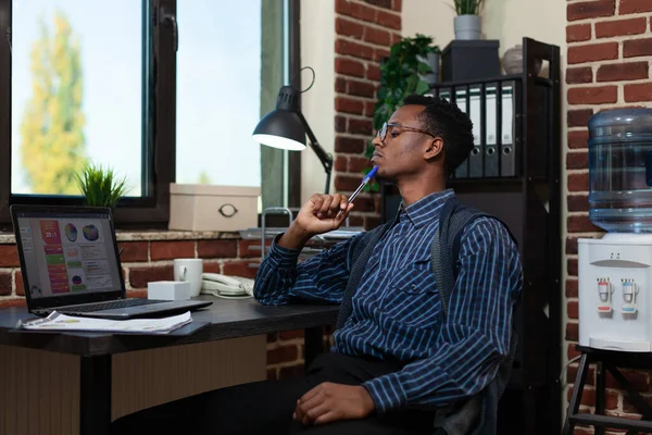 African american entrepreneur touching chin with pen looking focused at laptop screen — Stockfoto