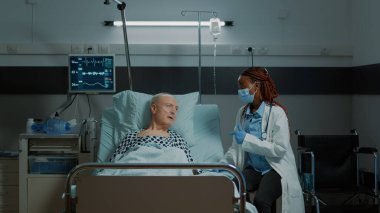 Afro-Amerikalı doktor hastane koğuşunda hasta bir hastayla konuşuyor.