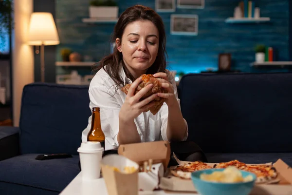 Frau sitzt auf Couch und schaut sich leckeres Burger-Abendessen im Wohnzimmer an — Stockfoto
