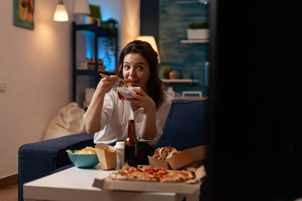 Happy woman enjoying delicious unhealthy takeaway asian noodles with chopsticks while watching television — Zdjęcie stockowe