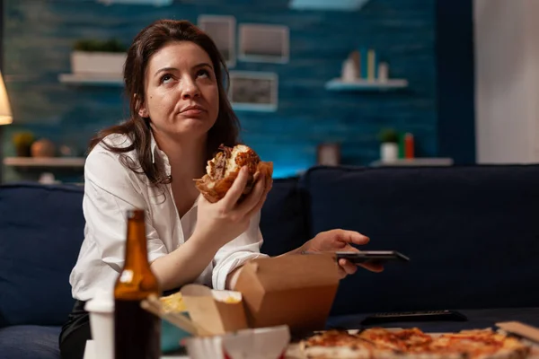 Sıkılmış bir kadın akıllı telefondan sosyal medyada geziniyor. Lezzetli hamburger yemekleri yiyor. — Stok fotoğraf