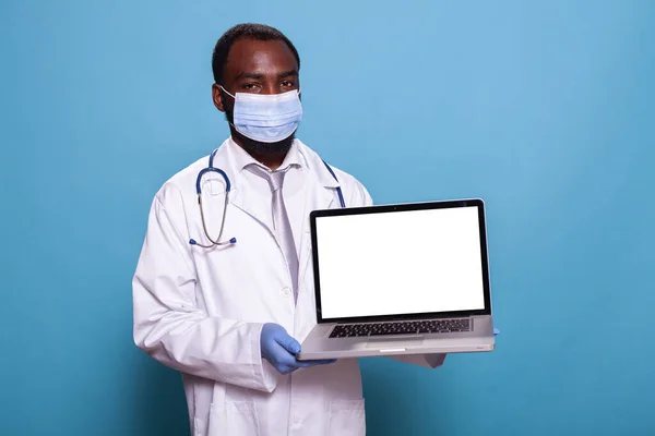 Medico indossare indumenti protettivi medici che presentano laptop con schermo bianco mockup — Foto Stock