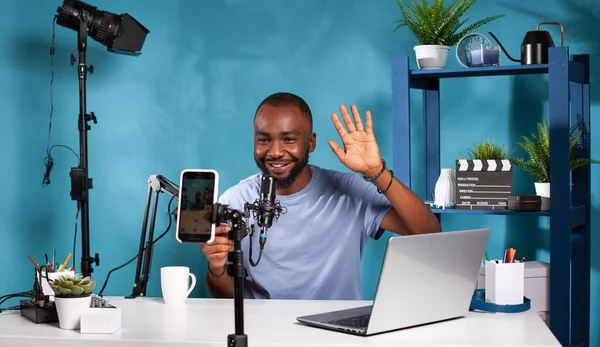 Vlogger sonriente saludando frente al teléfono inteligente de grabación sentado en el escritorio — Foto de Stock