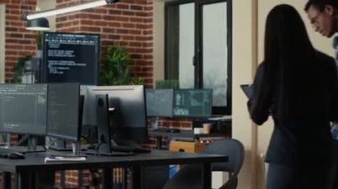 Two software developers holding laptop with coding interface walking towards desk and sitting down