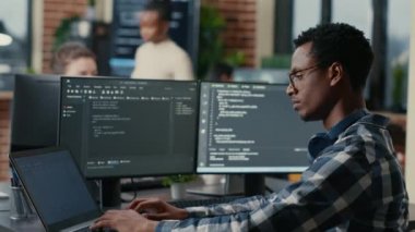 Sofware developer thinking while touching beard while typing on laptop sitting at desk with multiple screens