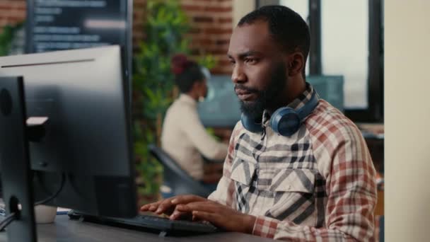 コンピュータ画面を見て働くワイヤレスヘッドフォンを身に着けているアフリカ系アメリカ人プログラマーの肖像画 — ストック動画