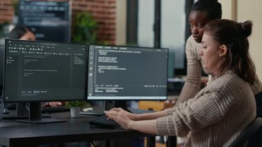 Junior programer writing algorithm under experienced colleague supervision sitting at desk with multiple screens