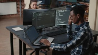 Portrait of tired programer rubbing eyes while typing on laptop sitting at desk with multiple screens parsing code