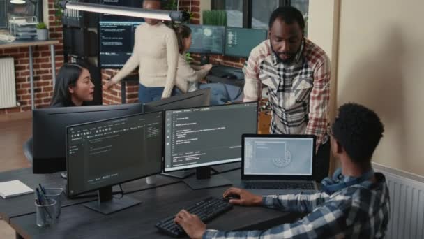 Programador sentado na mesa com várias telas executando código falando com colega — Vídeo de Stock