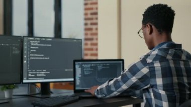 Focused software developer writing code on laptop looking at multiple screens with programming language