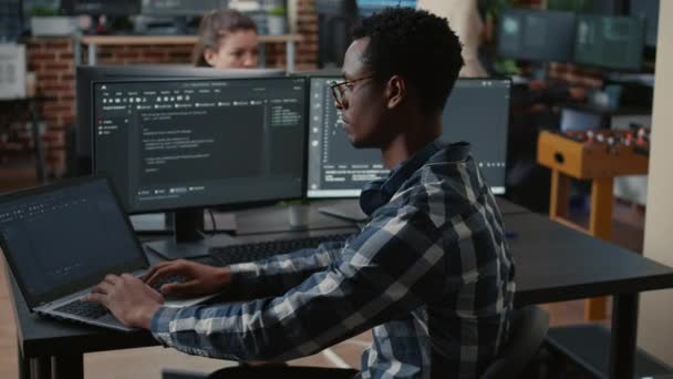 Retrato del programador afroamericano escribiendo en el ordenador portátil sentado en el escritorio con múltiples pantallas de análisis de código — Vídeos de Stock