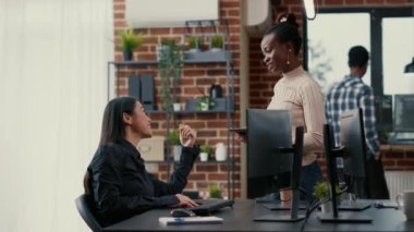 Programer standing up holding digital tablet talking with coder sitting at desk in front of computer screen