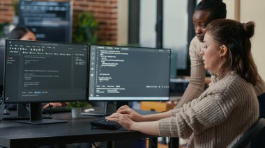 Junior programer writing algorithm under experienced colleague supervision sitting at desk with multiple screens