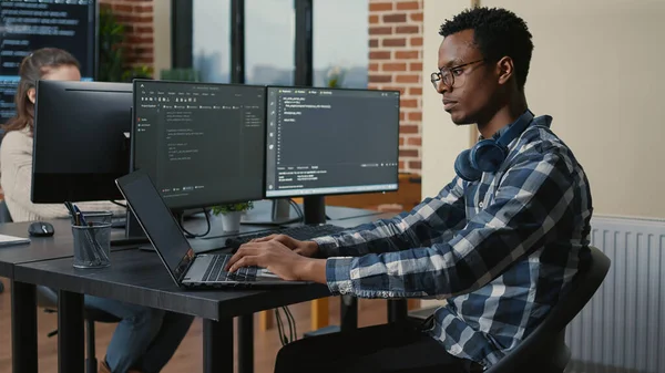 Programer thinking while touching beard and fixing glasses while typing on laptop sitting at desk — ストック写真