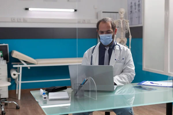 Man doctor wearing face mask and white coat using computer — 스톡 사진