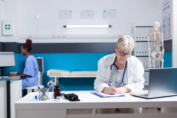 Portrait of senior medic taking notes on checkup papers at desk in office — Stockfoto
