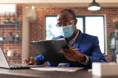 Employee analyzing clipboard files at desk to plan business project, wearing face mask