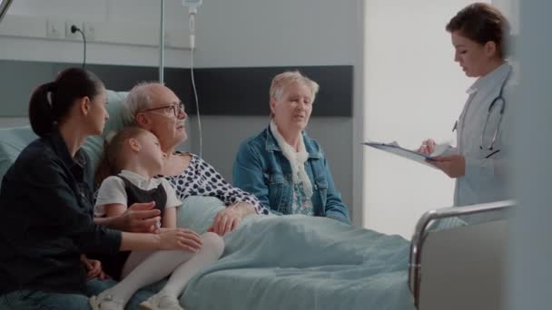 Лікар проводить огляд з сім'єю, яка відвідує хворого пенсіонера в лікарняному відділенні — стокове відео