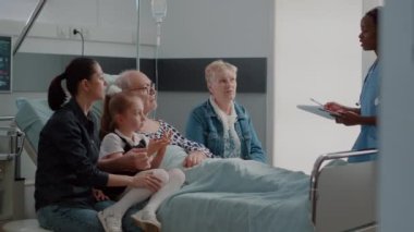 Aile, hastane koğuşunda emekli bir adamı ziyaret ediyor ve hemşireyle konuşuyor.