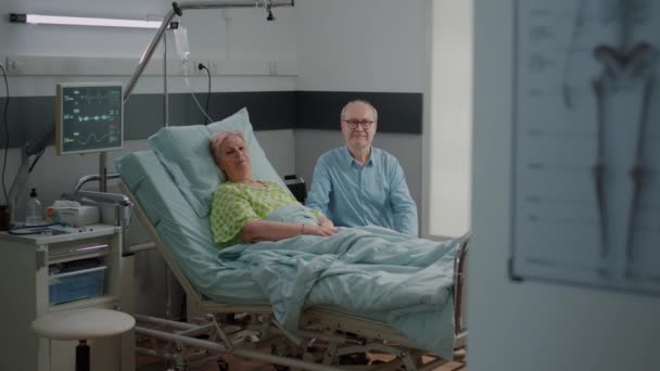 Portrait of pensioner with disease laying in bed and man giving assistance — Stock Video