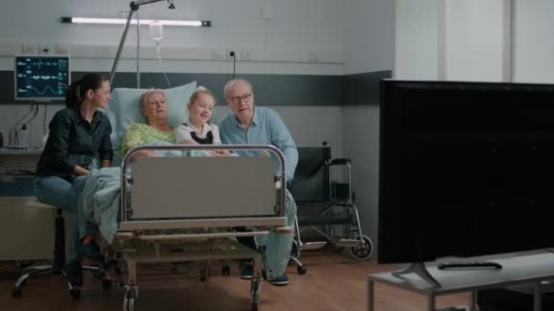 Family watching television and visiting senior patient in hospital ward — Αρχείο Βίντεο