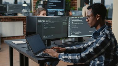Portrait of tired programer rubbing eyes while typing on laptop sitting at desk with multiple screens parsing code