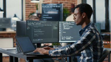 Portrait of african american developer using laptop to write code sitting at desk with multiple screens parsing algorithm