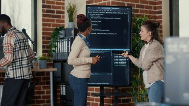 Programmer holding digital tablet analyzing code on wall screen tv while colleague is looking for errors