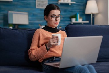 Student sitting on sofa holding cup and laptop.
