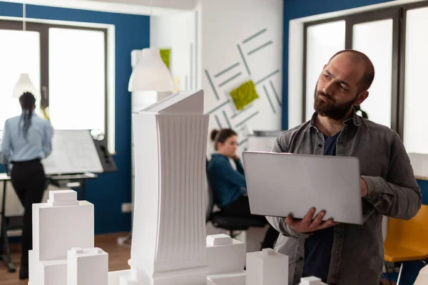Arkitektinspektör med laptop lutande huvud tittar på maquette — Stockfoto