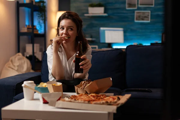 Mulher relaxante no sofá assistindo televisão enquanto bebe cerveja artesanal e come batatas fritas — Fotografia de Stock