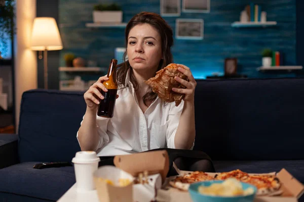 Mujer después del trabajo de oficina comiendo sabrosa hamburguesa para llevar sosteniendo botella de cerveza en el sofá viendo noticias — Foto de Stock