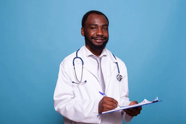 Médico sorridente confiante posando em jaleco branco com estetoscópio segurando prancheta — Fotografia de Stock