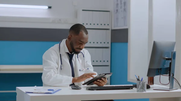 Médico terapeuta afroamericano sentado en el escritorio en la oficina del hospital sosteniendo la computadora tableta —  Fotos de Stock