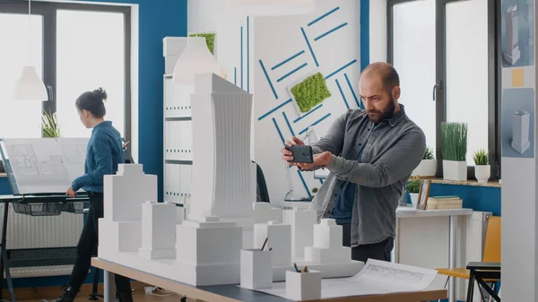 Людина-інженер використовує смартфон для роботи над будівельною моделлю та компонуванням в архітектурному офісі — стокове фото