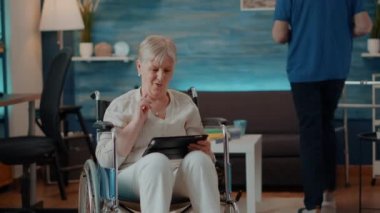 Tekerlekli sandalyede otururken dijital tablet kullanan yaşlı kadın.