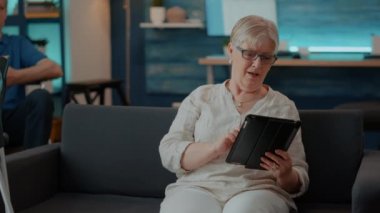 Oturma odasında dokunmatik ekranlı dijital tablet kullanan yaşlı kadın