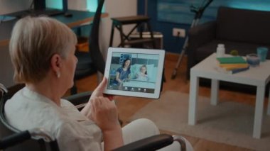 Tekerlekli sandalyedeki yaşlı kişi elinde online video çağrısı ile tablet tutuyor.