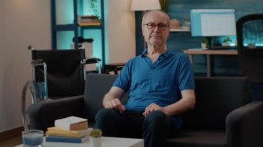 Evde tekerlekli sandalyenin yanında oturan yaşlı kişinin portresi.