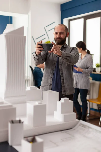Arquitecto de diseño sonriente sosteniendo smartphone para fotografiar escritorio con modelo arquitectónico —  Fotos de Stock