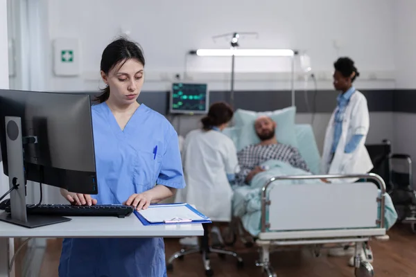 护士在检查有病史的剪贴板时，使用个人电脑填写病人入院表 — 图库照片