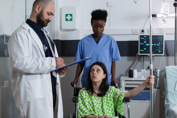 Enfermeira assistente mulher com doença sentado em cadeira de rodas segurando pólo intravenoso enquanto médico segurando prancheta — Fotografia de Stock