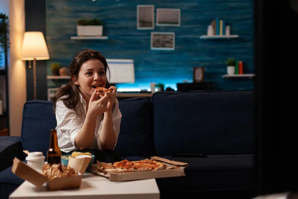 Mosolygó nő eszik egy szelet forró pizza szállítás ül a kanapén nézi a családi show a televízióban — Stock Fotó