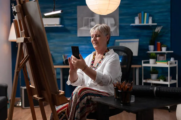 Artista idoso sentado na frente do cavalete com tela de navegação smartphone — Fotografia de Stock