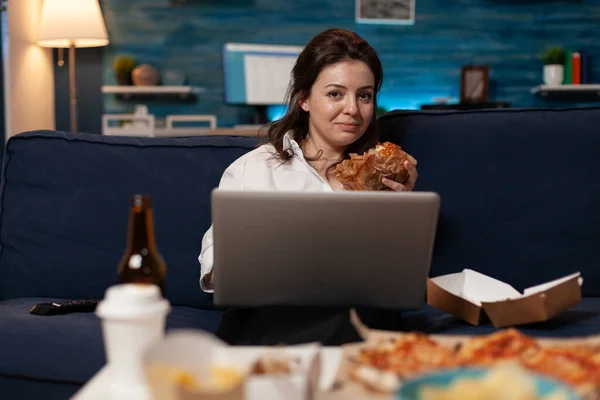 Programador relajante en sofá sosteniendo portátil comiendo una sabrosa hamburguesa para llevar y menú de entrega de comida rápida — Foto de Stock