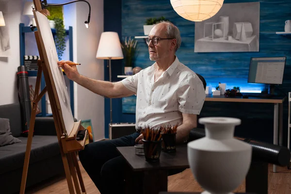 Profesor de arte sénior dibujando jarrón sobre lienzo de papel en caballete usando un lápiz afilado sentado en la mesa —  Fotos de Stock