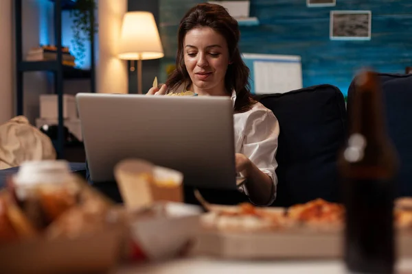 Mujer navegando por las redes sociales en el ordenador portátil mientras tiene bocadillo de comida rápida sentado en el sofá — Foto de Stock