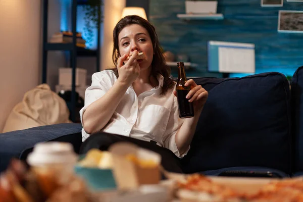 Kobieta jedząc kawałek gorącej dostawy pizzy siedzi na kanapie trzymając butelkę piwa patrząc na telewizję — Zdjęcie stockowe